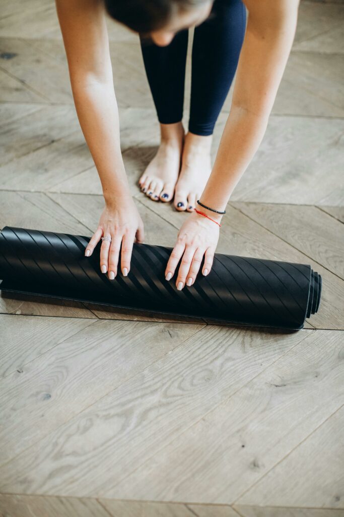 Close-up of a woman rolling a black yoga mat on a wooden floor, focusing on fitness and wellness.